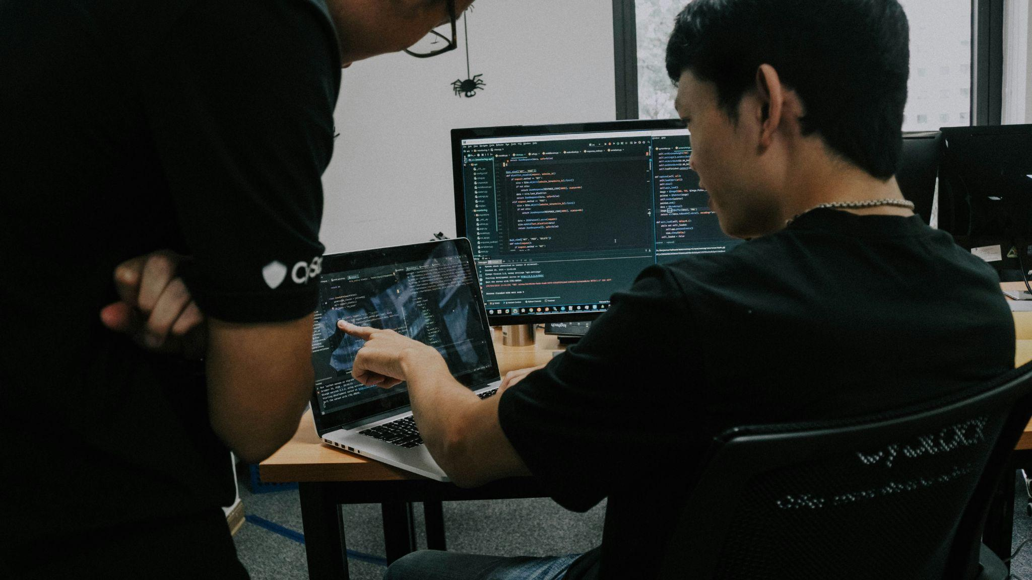 Two developers in an office reviewing quantum code together on a laptop and a monitor full of Python source.