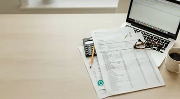 Desk with laptop, calculator, pencil, glasses and coffee on printed cost spreadsheets, illustrating AI agent project budgeting and timeline planning.