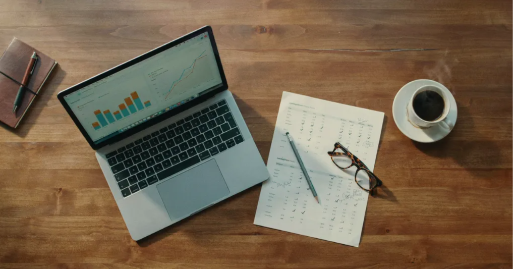 Flat-lay of a laptop showing analytics beside a checklist, glasses and coffee — evaluating software costs.