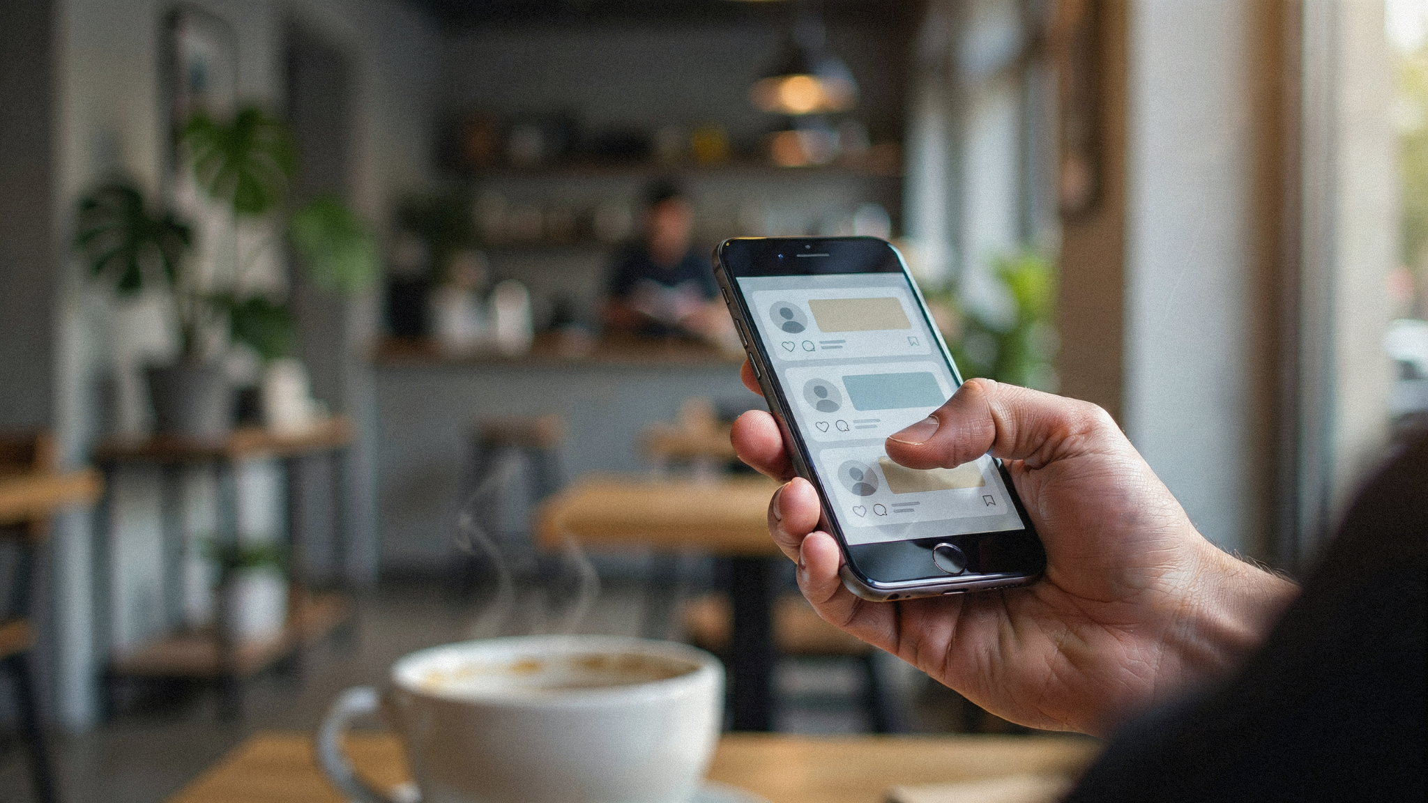 Hand holding a phone showing a social media feed in a cafe.