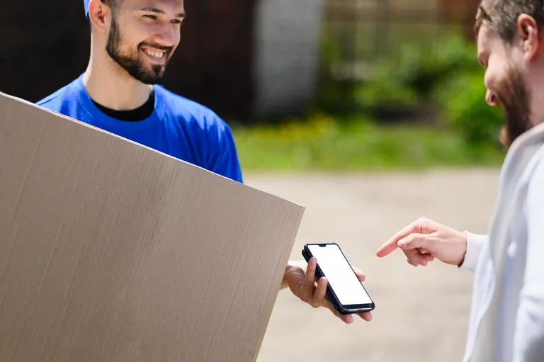 A courier in a blue cap and blue T-shirt stands outdoors holding a large cardboard box against his shoulder, smiling at the person in front of him. In his other hand he holds out a smartphone with a blank white screen, and the person, in a white sweater, reaches in and points a finger at it. Behind them a paved driveway and blurred green foliage.